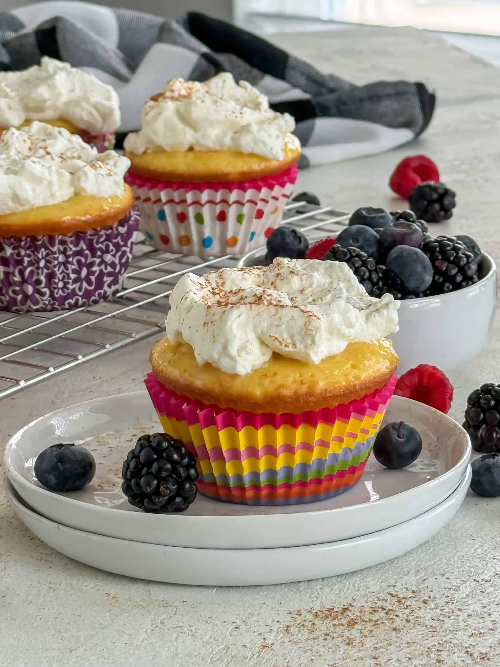 A colorful cupcake topped with whipped cream and a dusting of cinnamon, placed on a white plate with fresh berries.