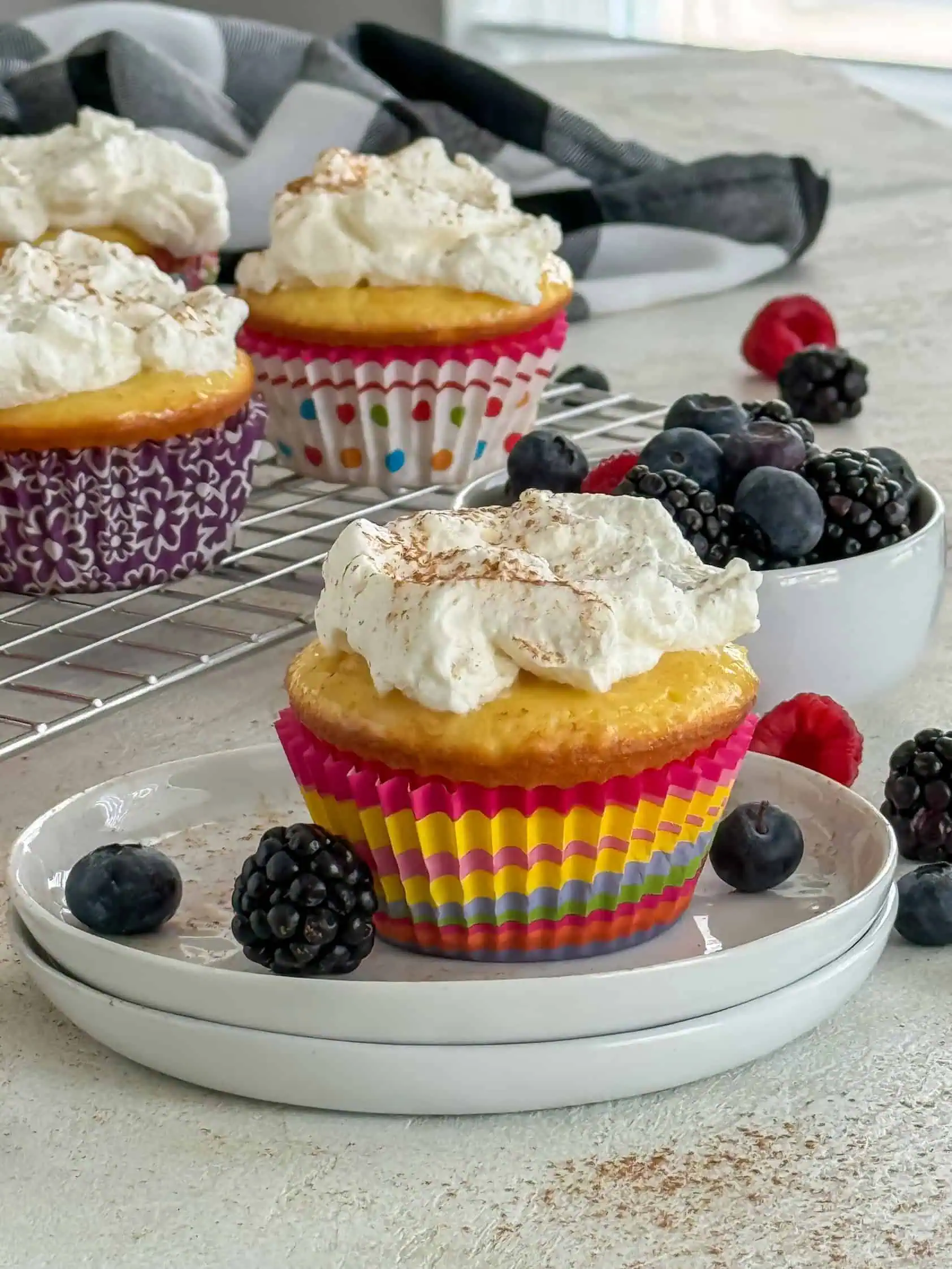 A colorful cupcake topped with whipped cream and a dusting of cinnamon, placed on a white plate with fresh berries.