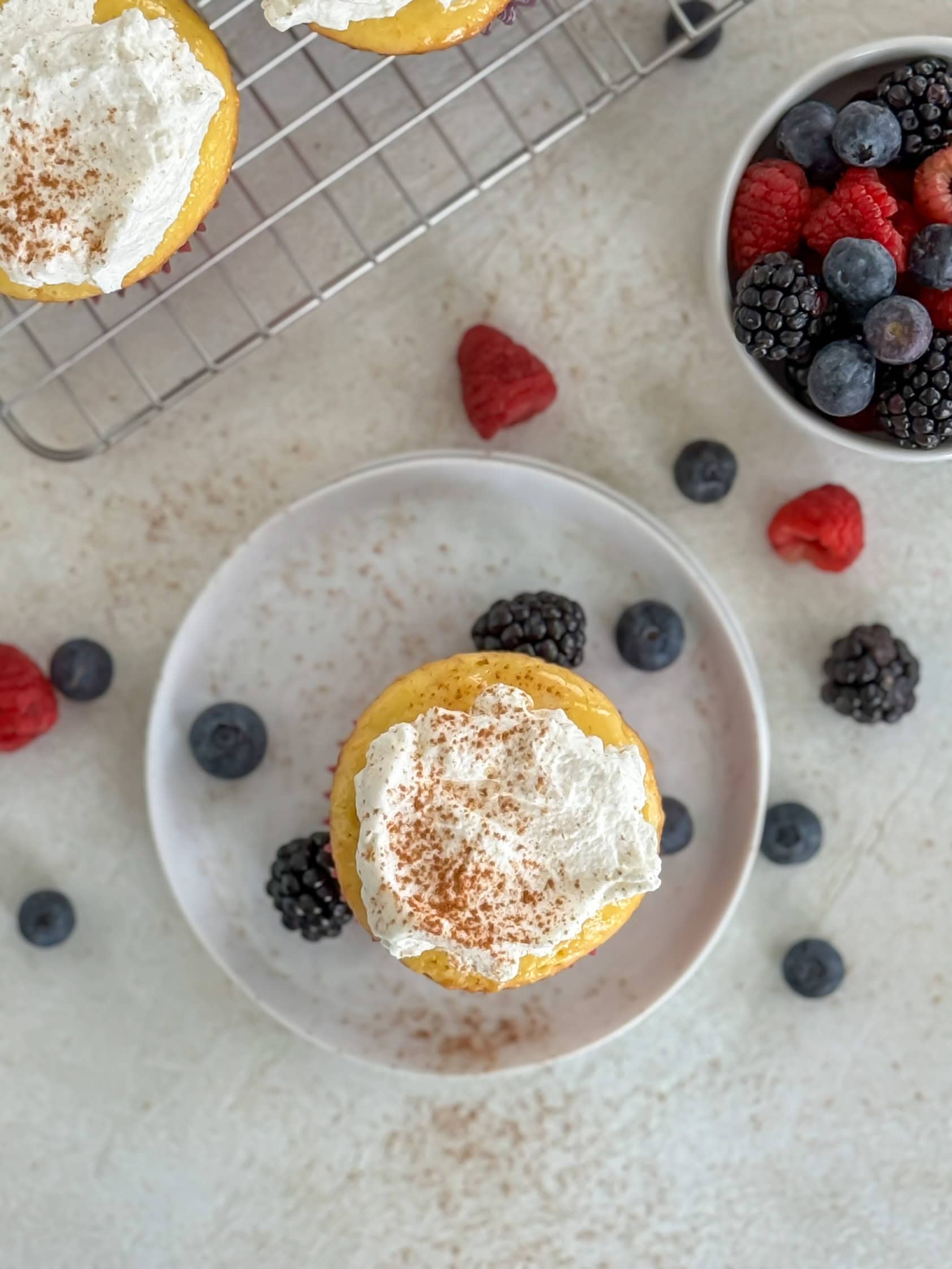 An overhead shot of a cupcake topped with whipped cream and a dusting of cinnamon on a white plate, surrounded by scattered fresh berries.