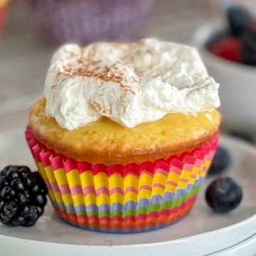 A single cupcake in a colorful liner, topped with whipped cream and a dusting of cinnamon, with a fresh blackberry and blueberry on the plate.