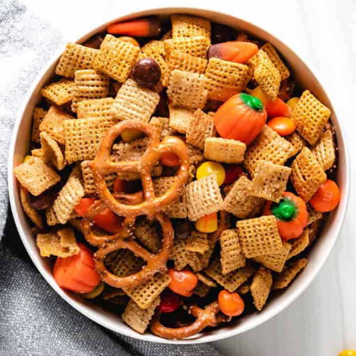 A bowl filled with halloween snack mix, featuring a combination of corn chex, rice chex, mini pretzels, autumn mix candy, reese's pieces, and m&m's candy in harvest colors.