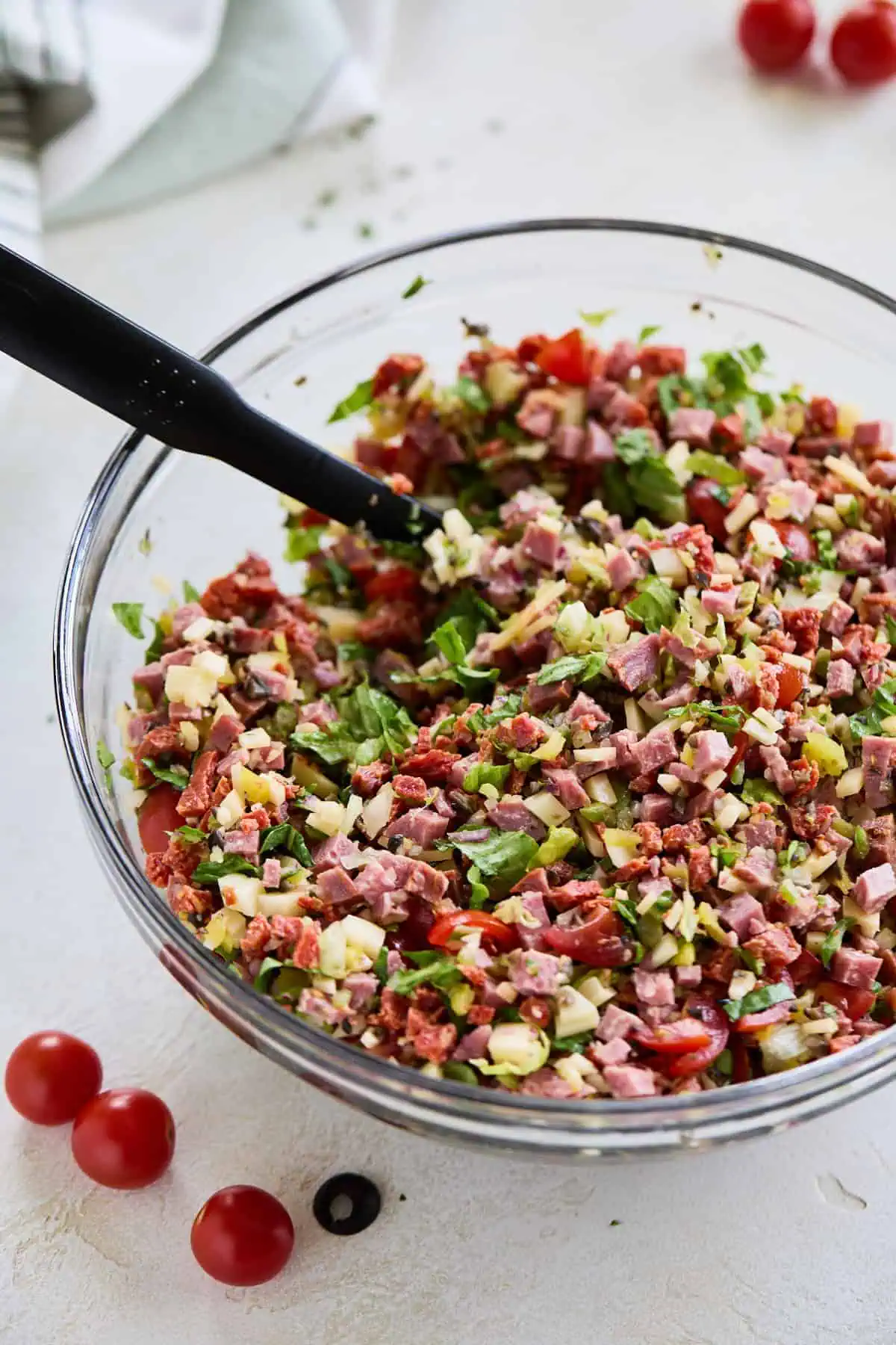 A large glass bowl showing a mix of diced italian meats, cheese, and chopped vegetables being combined with the creamy dressing.