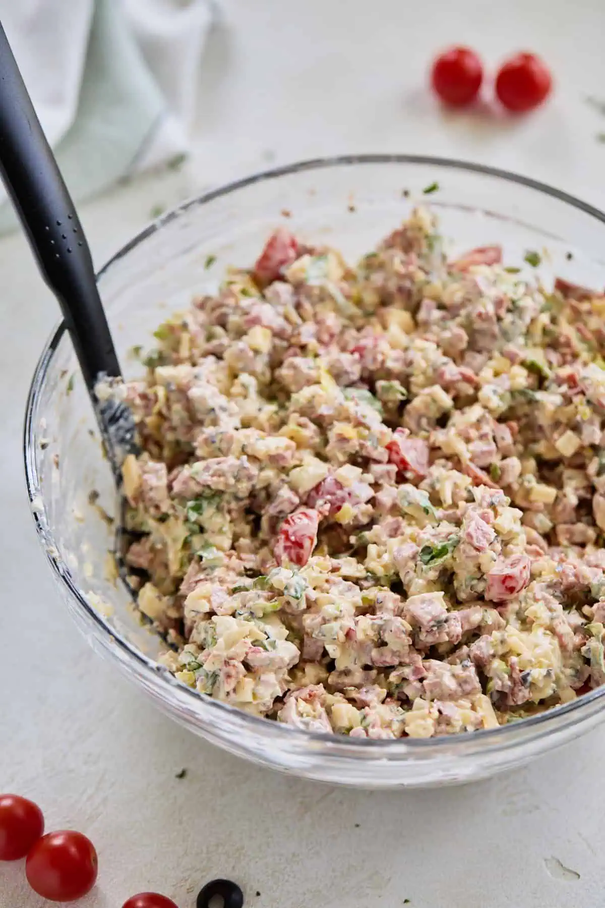 A large glass bowl filled with a colorful mixture of diced italian meats, cheese, vegetables, and lettuce, being stirred together with a black spoon.