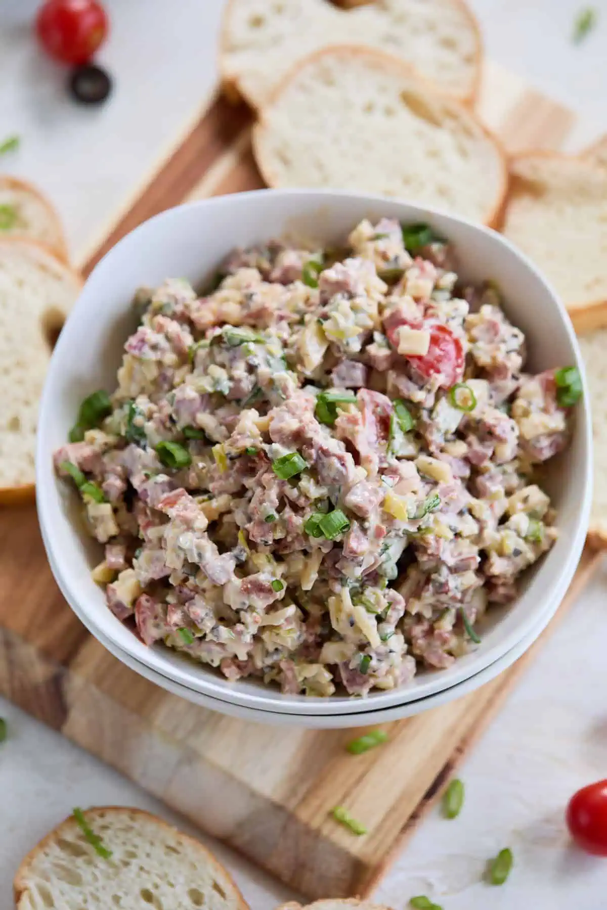 A white bowl of hoagie dip on a wooden board, with slices of french baguette around it, showcasing the colorful and creamy mixture of italian meats, cheese, and vegetables.