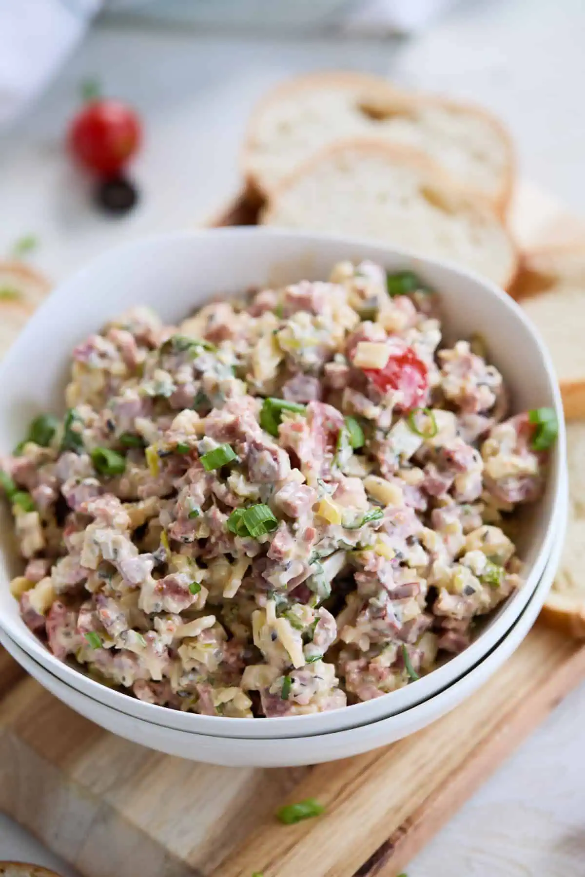 A white bowl of hoagie dip on a wooden board, with slices of french baguette around it, showcasing the colorful and creamy mixture of italian meats, cheese, and vegetables.