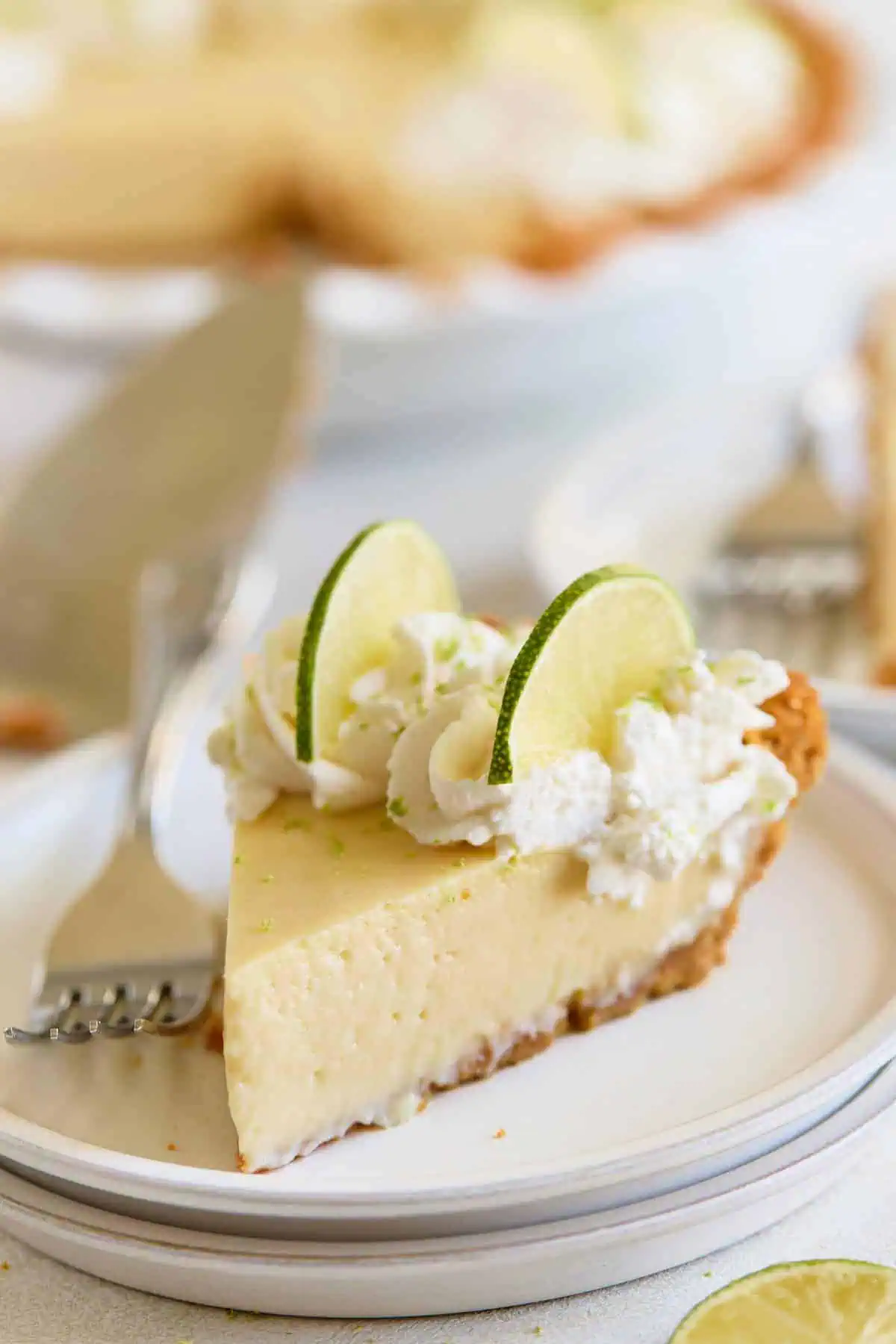 A close-up slice of creamy pie topped with whipped cream and lime slices on a white plate, with the whole pie in the background.