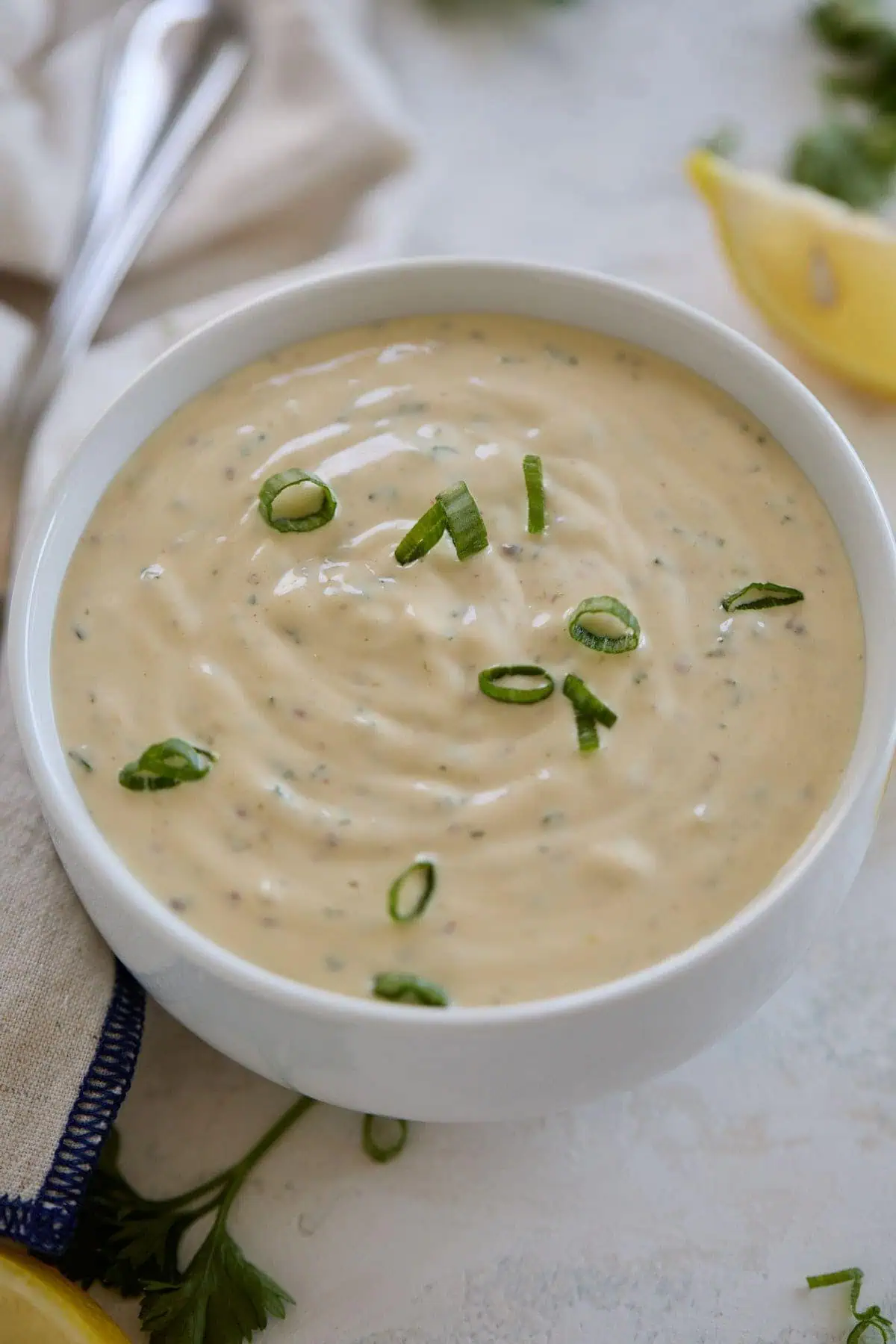 Close-up of remoulade sauce in a bowl, garnished with green onions, with a blurred background of lemon wedges and napkin.