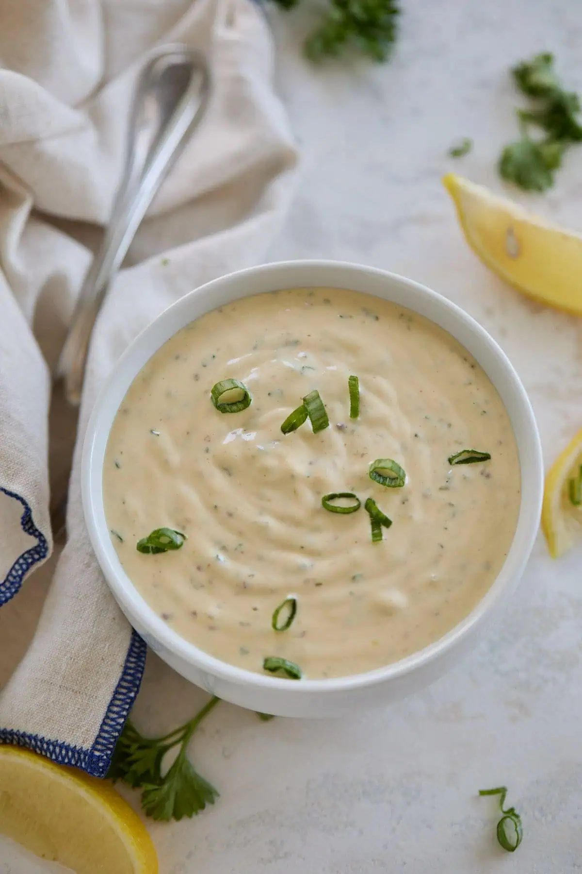 Overhead view of remoulade sauce in a bowl, garnished with green onions, with a cloth napkin and lemon wedges around.