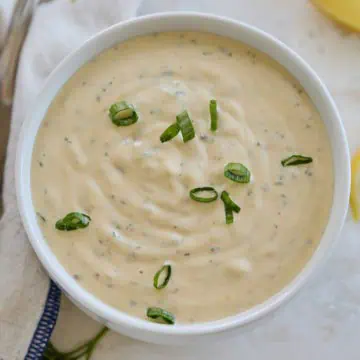 Close-up of remoulade sauce in a bowl, garnished with green onions, with parsley and lemon wedges in the background.