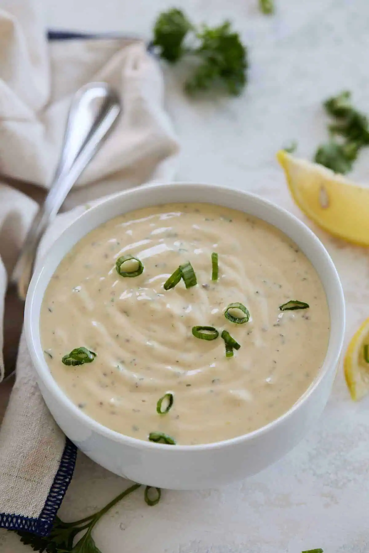 A bowl of creamy dipping sauce garnished with green onions, placed on a white surface with a cloth napkin beside it.