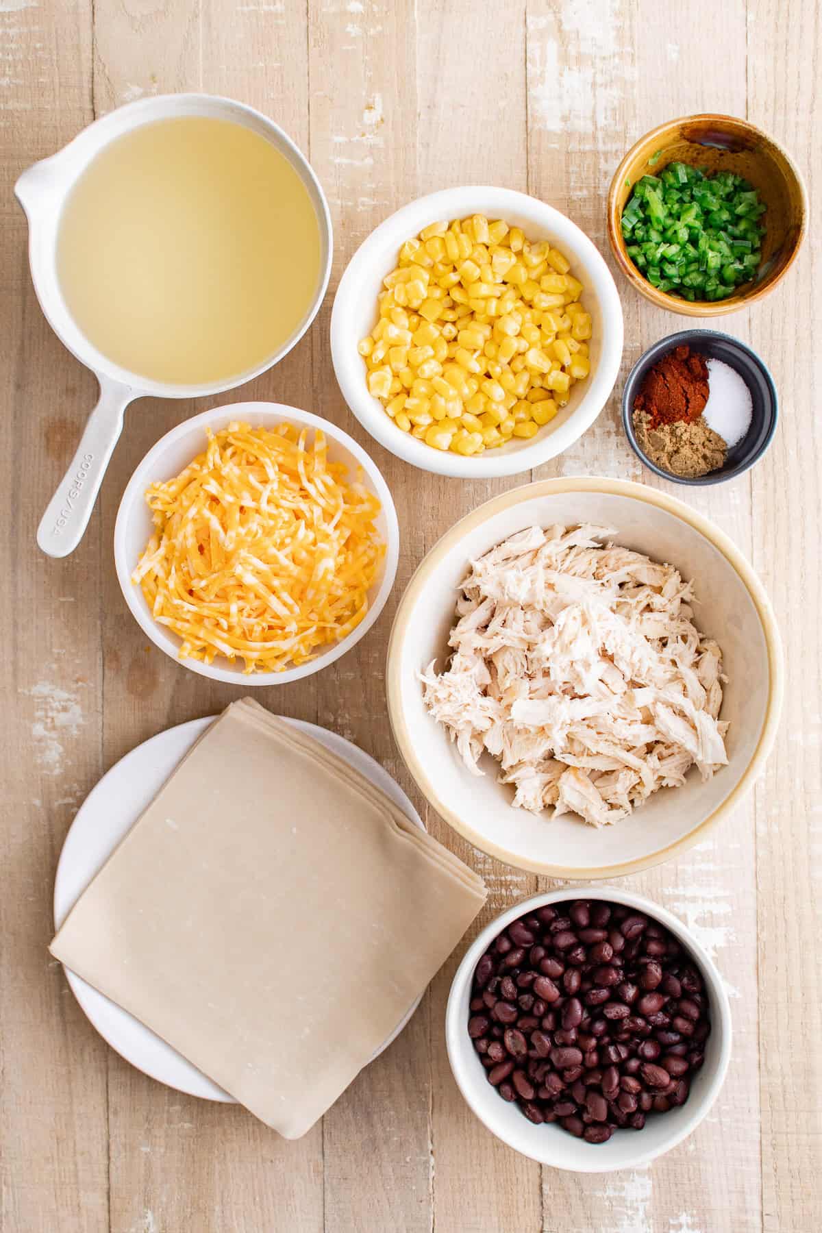 Vegetable oil in a measuring cup, shredded chicken, shredded colby jack cheese, black beans, thawed corn, finely minced jalapeño, ground cumin, chili powder, salt, and egg roll wrappers arranged on a wooden surface.