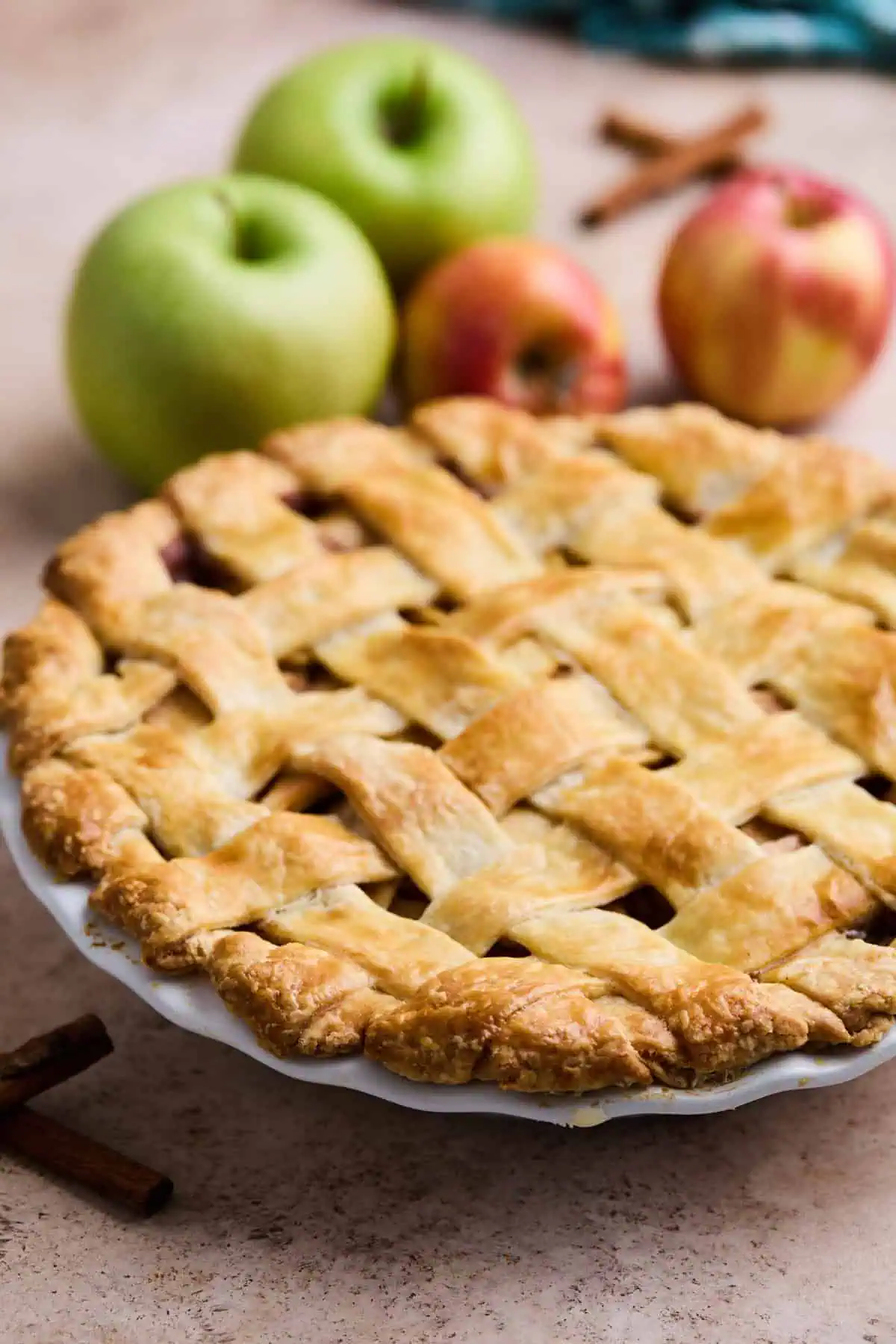 A whole lattice-topped apple pie with a golden-brown crust, with fresh apples and cinnamon sticks in the background.