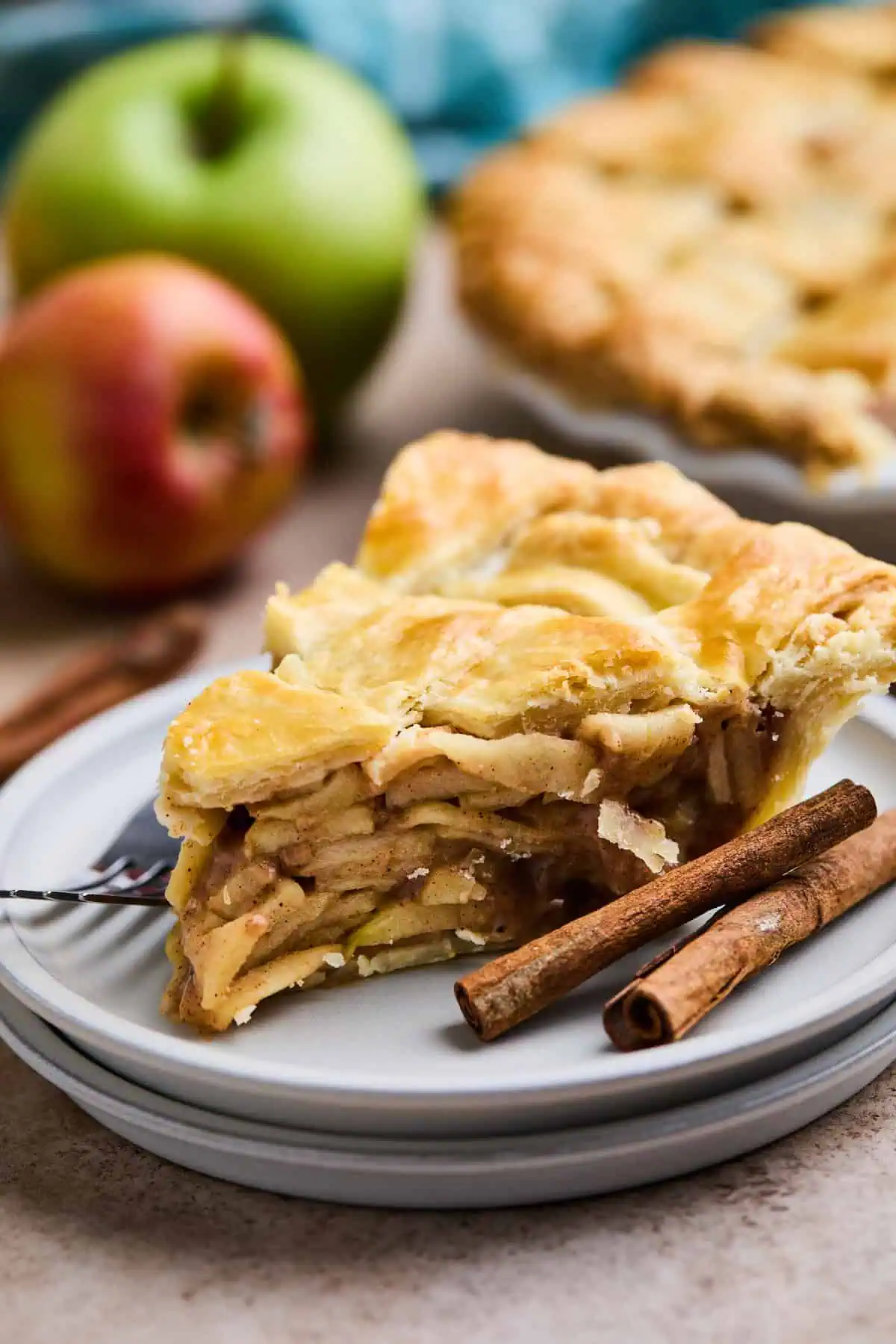 A slice of apple pie with a flaky, golden crust on a plate, with cinnamon sticks and whole apples blurred in the background.