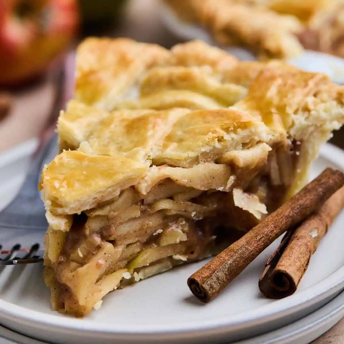 A close-up slice of apple pie with a golden crust, showcasing the layered apple filling, with cinnamon sticks and a fork beside it.