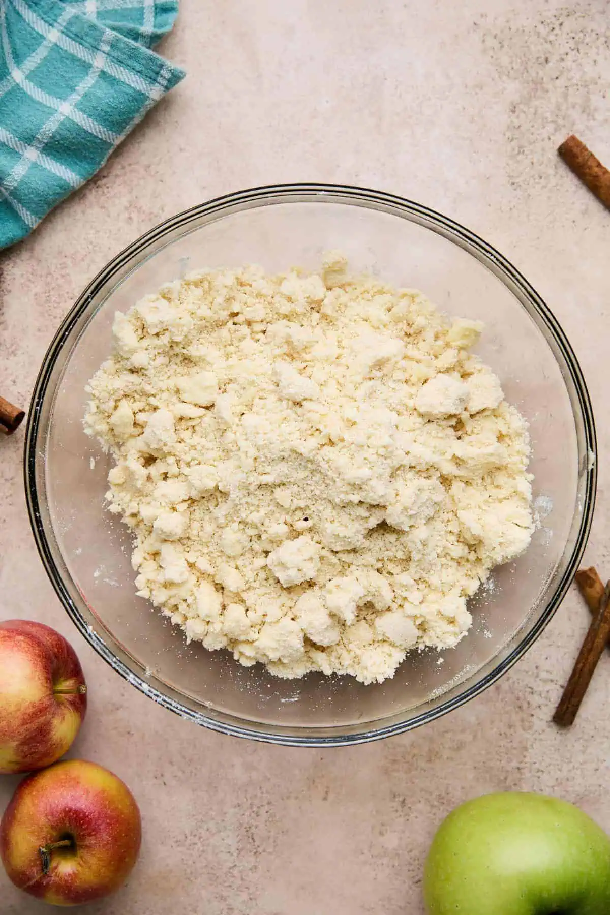 A glass bowl containing a crumbly mixture, likely a dough or topping, with apples and cinnamon sticks around.