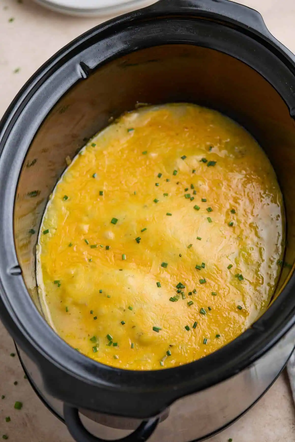 Crockpot filled with creamy ranch chicken topped with melted cheddar cheese and garnished with chopped chives.