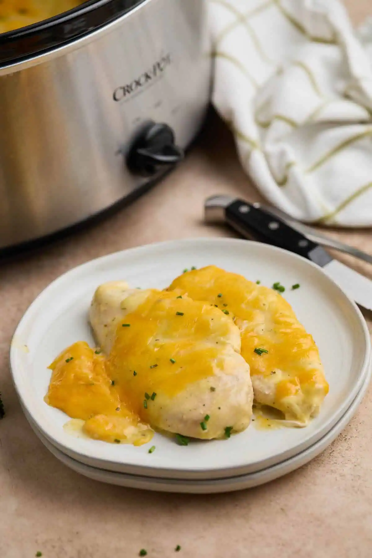 Creamy ranch chicken breast on a white plate, with melted cheddar cheese and a sprinkle of chives, served next to a crockpot.