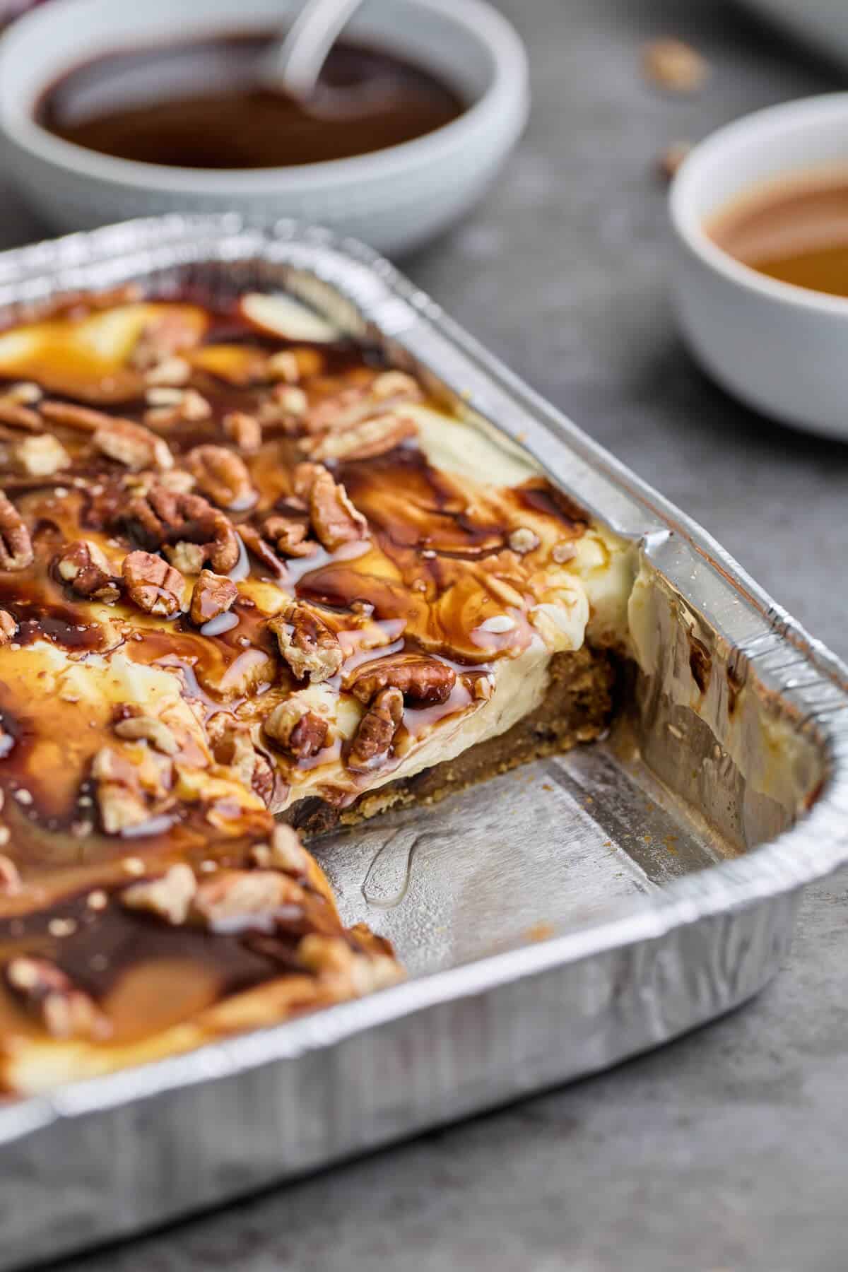 A close-up of turtle cheesecake with caramel and chocolate drizzles and pecans, showing a slice cut out.