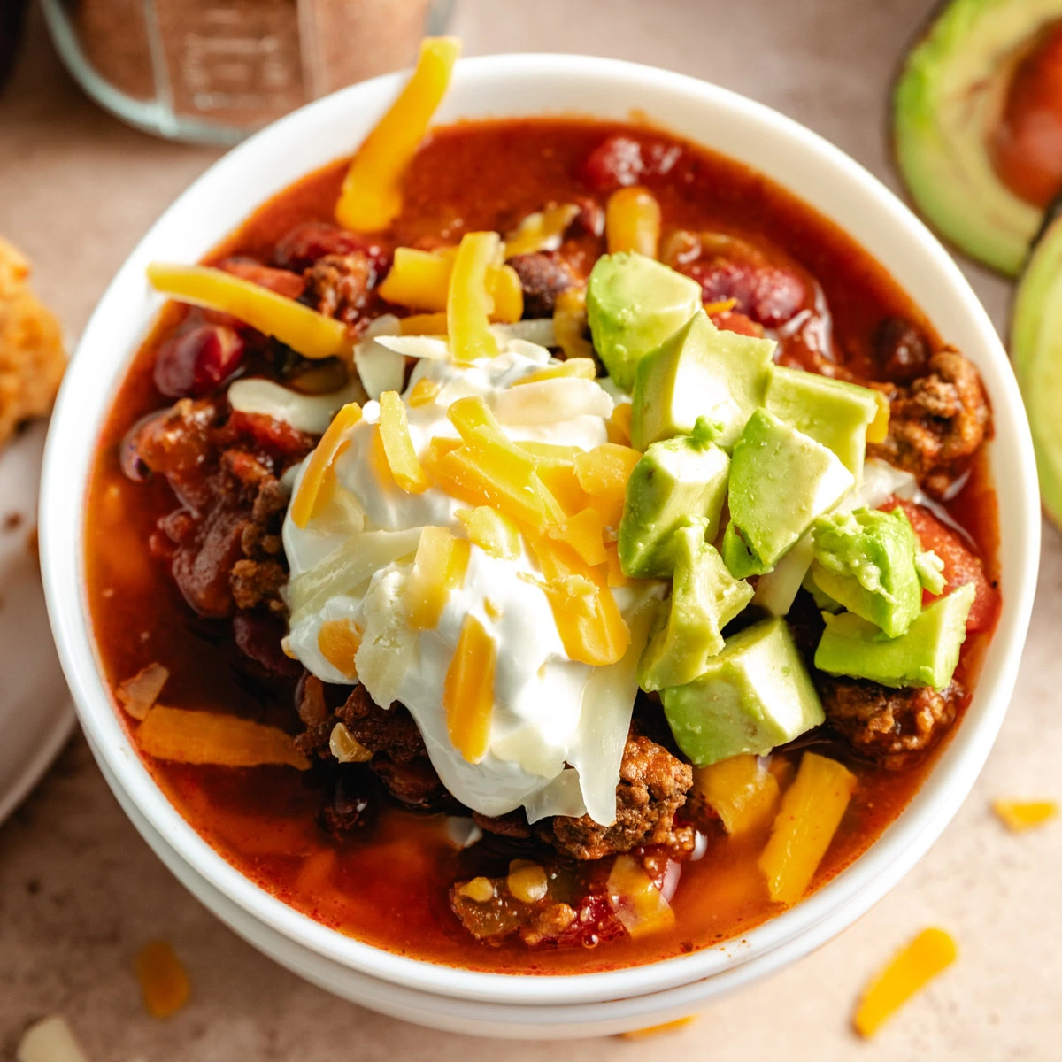 A bowl of chili topped with sour cream, shredded cheese, and diced avocado.