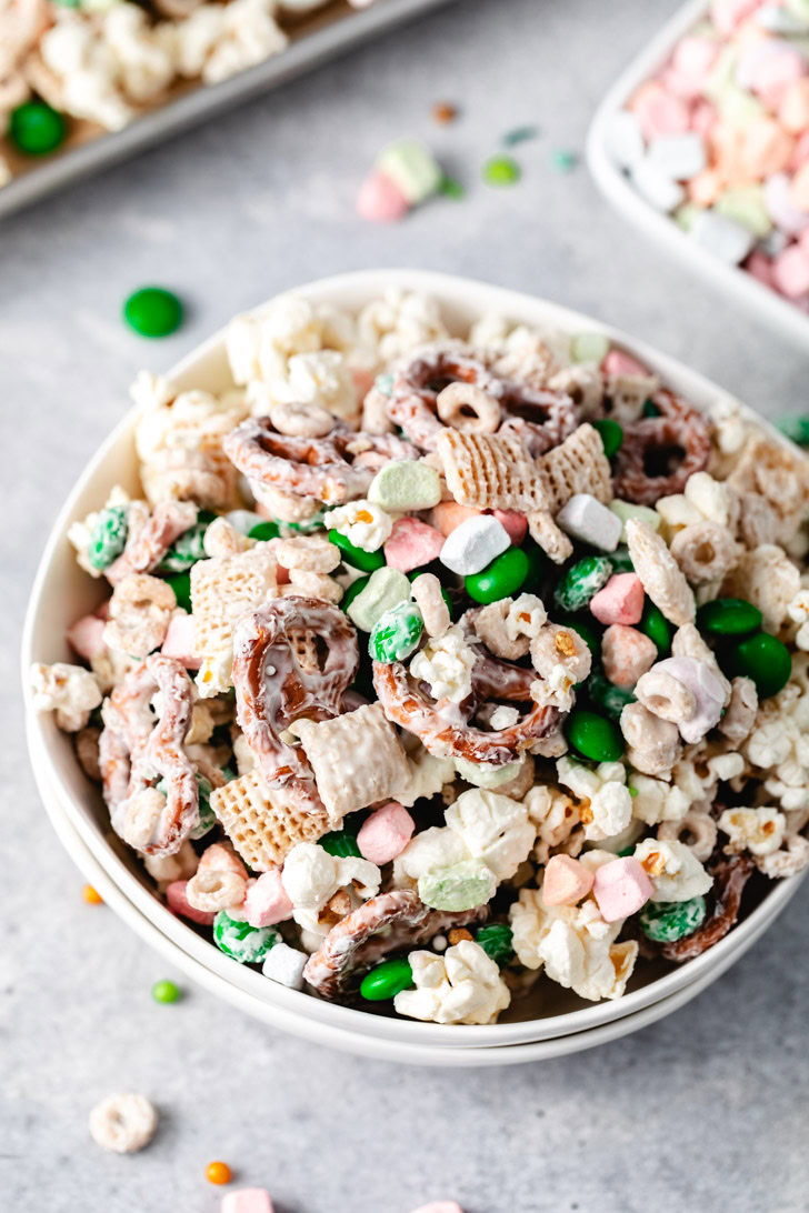 A close-up of a bowl of leprechaun bait snack mix with green m&ms, cereal, and pretzels covered in vanilla melting wafers.